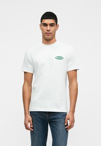 Balta medvilnės marškinėliai su trumpomis rankovėmis, apvalia kaklo iškirpte ir žaliu logotipo užsklanda kairėje krūtinės pusėje. Dėvimi su tamsiai mėlynomis džinsinėmis kelnėmis.