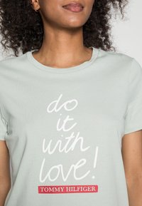 Mujer vistiendo una camiseta verde clara con el texto blanco "¡hazlo con amor!" y un parche rojo con el logo de "Tommy Hilfiger".