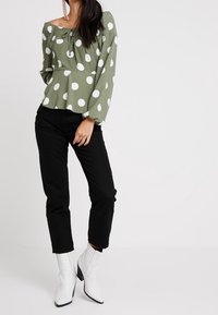 Blusa verde oliva con pois bianchi, design off-shoulder, maniche lunghe, vita cintrata, abbinata a pantaloni neri e stivaletti bianchi.