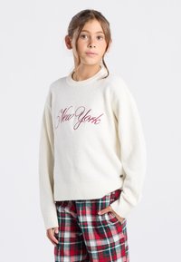 Pull crème avec "New York" brodé en bordeaux, col et poignets côtelés, associé à un pantalon à carreaux rouge, vert et blanc.