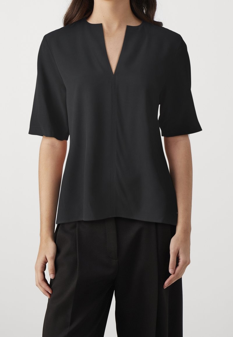 Femme portant un haut noir à manches courtes avec un col en V entaillé et un pantalon noir, debout devant un fond uni.