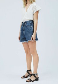 Blusa bianca con dettagli in pizzo, pantaloncini di jeans a vita alta con orlo sfilacciato e sandali neri con borchie e tacco spesso.