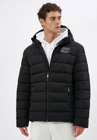 Sort pufferjakke med hætte, lynlåslommer, struktureret quiltet mønster og hvid broderet logo. Båret over en hvid hoodie.