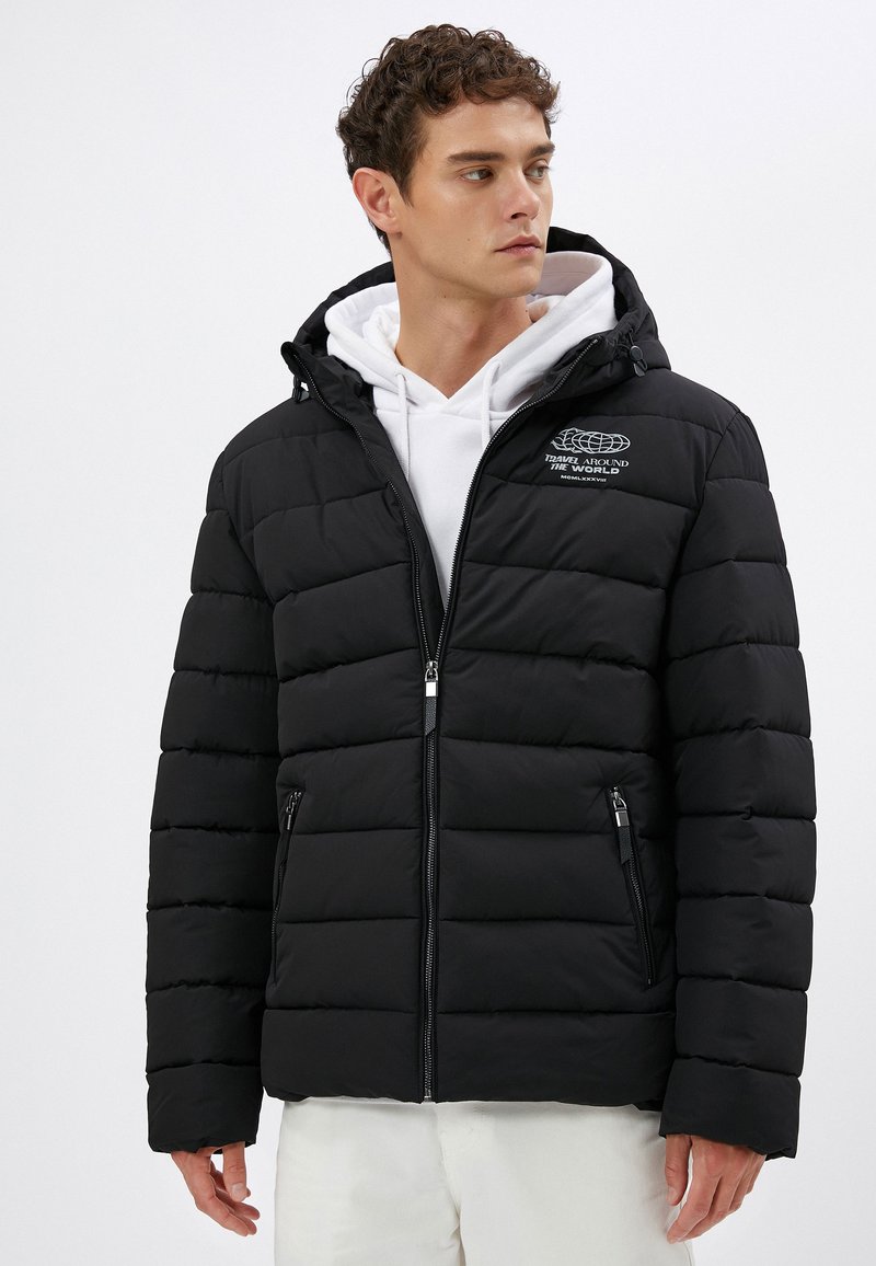 Sort pufferjakke med hætte, lynlåslommer, struktureret quiltet mønster og hvid broderet logo. Båret over en hvid hoodie.