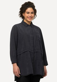 Camisetta nera a maniche lunghe con collo button-down, dotata di taschino sul petto e orlo curvo per un design stratificato. Texture morbida e liscia.