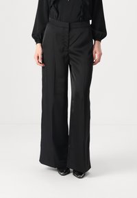 Pantaloni neri a gamba larga in tessuto morbido con un design sartoriale, featuring una vita alta e dettagli minimi. Abbinati a scarpe con punta.