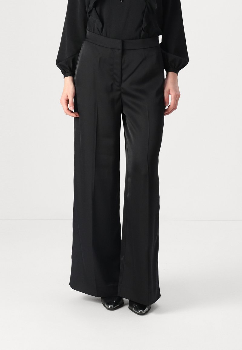 Pantaloni neri a gamba larga in tessuto morbido con un design sartoriale, featuring una vita alta e dettagli minimi. Abbinati a scarpe con punta.