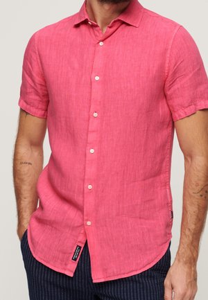 Mand iført en skarp pink kortærmet skjorte med krave og marineblå bukser med striber, hænderne i lommerne.