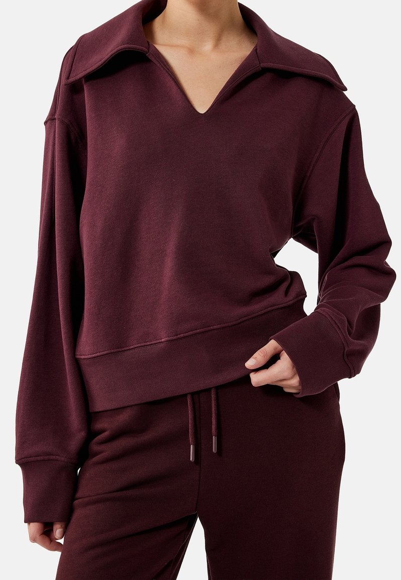 Femme portant un pull bordeaux foncé à col large et un pantalon assorti à cordon, main tenant l'ourlet du pull.