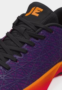 Nahaufnahme eines lila Sportschuhs mit schwarzen Schnürsenkeln, orange-rot gefärbter Sohle, strukturiertem geometrischem Muster und orangefarbenem Logo auf schwarzer Zunge.