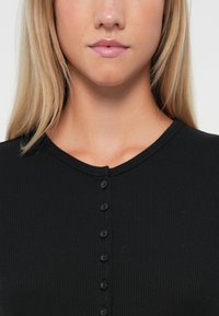 Svart, stripete langermet topp med rund hals og en rad av seks svarte knapper foran. Glatt tekstur, tettsittende design.