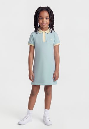 Fiatal lány áll, világoskék, bézs galléros és mandzsettás pólóruhában, fehér zokniban és fehér tornacipőben, egyszínű háttér előtt.