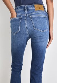 Los vaqueros azules, hechos de mezclilla, presentan un diseño deslavado con cinco bolsillos, un parche de cuero marrón y un corte recto.