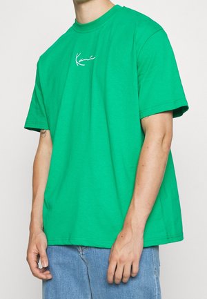 Person iført en skriggrøn kortærmet t-shirt med hvid skrift på brystet, kombineret med blå jeans, stående mod en ensfarvet baggrund.