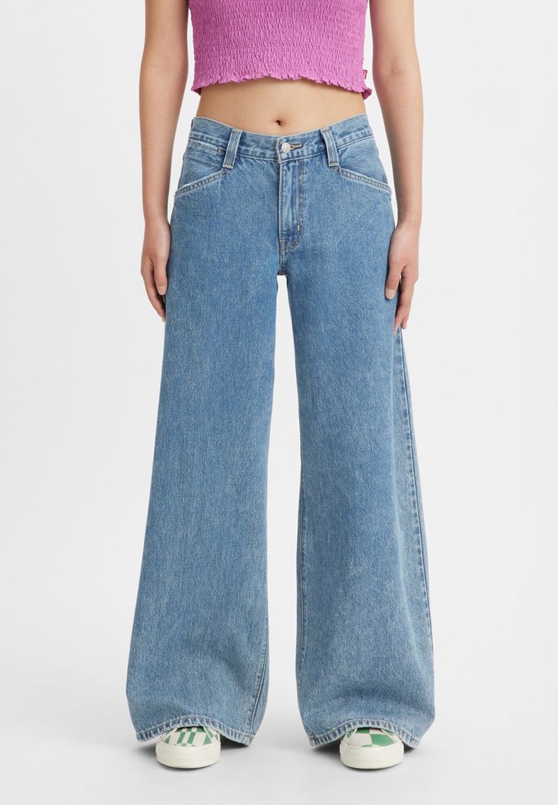 Personne portant un jean large bleu clair, des baskets blanches avec un motif vert et un top court rose texturé, debout devant un fond uni.