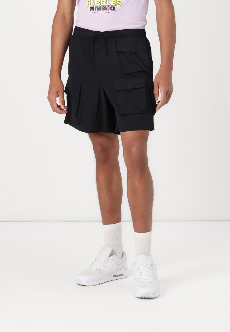 Black cargo shorts with multiple side pockets, waistband with drawstring, above-knee length, smooth fabric, paired with white sneakers and socks.