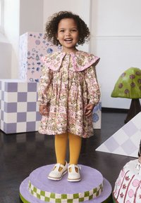 Ružové šaty s kvetinovým vzorom a veľkým volánovým golierom, v kombinácii so žltými pančuchami a bielymi topánkami Mary Jane, stojace na kruhovom podstavci.