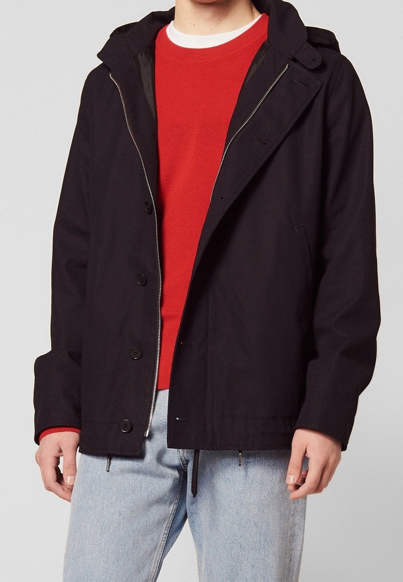 Czarna, lekka kurtka z kapturem, z przodu na zamek błyskawiczny, z zatrzaskami i kieszeniami po bokach, noszona na czerwonym swetrze, zestawiona z jasnoniebieskimi dżinsami.