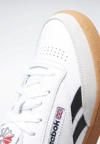 Bílé kožené tenisky s černými akcenty, šedým semišem a gumovou podrážkou. Obsahují perforace a logovou visačku s vlajkou Unie.