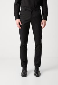 Pantalons slim noirs en tissu lisse, dotés de passants de ceinture, de poches latérales et d'un design classique. Portés avec des chaussures noires à lacets.