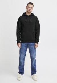 Black hoodie with "TOKYO" printed on the chest, featuring a front pocket and drawstring hood. Paired with light blue jeans and white sneakers.