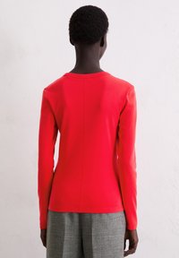 Czerwona bluzka z długim rękawem wykonana z gładkiej, lekkiej tkaniny, z zaokrąglonym dekoltem i prostym szwem z tyłu, zestawiona z szarymi spodniami.