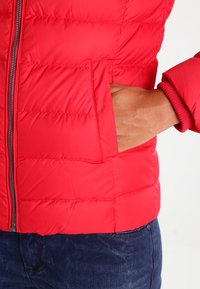 Czerwona, puchowa kurtka z pionowym wzorem pikowania, zapinana na zamek błyskawiczny i posiadająca kieszenie po bokach. Charakteryzuje się ściągaczami przy mankietach, wykonana z błyszczącego, lekkiego materiału.