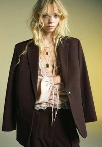 Blazer oversize marron associé à un haut rose clair avec des bordures en dentelle. Les accessoires comprennent des colliers en or superposés. Arrière-plan avec un dégradé doux.