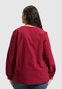 Rote Bluse mit Struktur, langen Ärmeln, rundem Ausschnitt und geknöpften Manschetten, von hinten vor neutralem Hintergrund gezeigt.