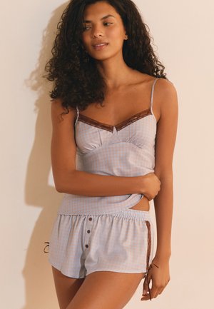 Vrouw met krullend haar die een lichtblauw geruit hemdje en bijpassende shorts met bruine kanten rand draagt, staand tegen een effen muur.
