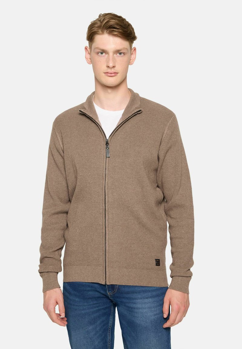 Brun zip-up cardigan i strukturerad stickning; har en ståkrage och svart dragkedja. Matchad med blå jeans och en enkel vit t-shirt under.