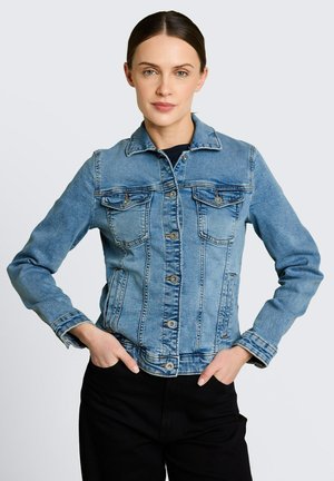 Frau mit glatten braunen Haaren trägt eine geknöpfte blaue Jeansjacke und schwarze Hose, steht mit Händen in den Taschen vor schlichten Hintergrund.