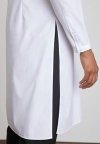 Camisa blanca de manga larga con una abertura lateral que revela un panel negro. La tela parece lisa con puños abotonados.