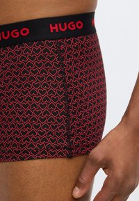 Cuecas boxers vermelhas e pretas com padrão de corações, apresentando uma cintura elástica preta com a inscrição "HUGO" em letras vermelhas. Textura suave.