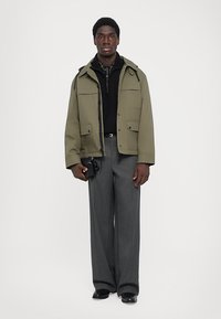 Mann trägt eine olivgrüne Jacke, einen schwarzen Reißverschluss-Pullover, graue weite Hosen, schwarze Schuhe und hält eine kleine schwarze Tasche.