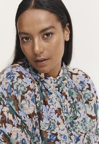 Kwiatowa bluzka w brązowych, niebieskich i zielonych kolorach, z zapięciem na guziki z przodu, wykonaną z miękkiego materiału i luźnym kroju z delikatnym wzorem.