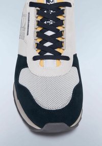 Sneaker caratterizzata da una combinazione di tela crema, suede nera e accenti gialli. La rete testurizzata e i lacci scuri completano il design sportivo.