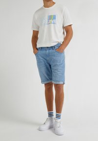 Ljusblå denimshorts med uppvikta mudd, matchade med en vit t-shirt med en färgglad "Lee Original" grafik. Vita sneakers och randiga strumpor.