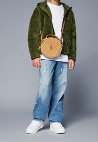 Chaquetón verde de felpa con cremallera, camiseta blanca, vaqueros azul holgados con manchas de pintura y zapatillas blancas. Bolso bandolera de pana redondo con logo.