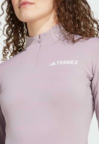 Maglietta lunga a maniche viola chiaro con colletto alto e mezza zip, che presenta una leggera texture sui lati e un logo TERREX bianco.