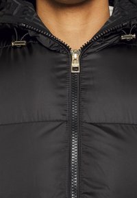 Zbliżenie czarnej kurtki puchowej z złotym zamkiem i stoperami sznurków przy kołnierzu, częściowo zapiętej na szyi osoby.