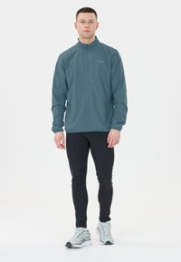 Teal lättvikts träningsjacka med hög krage, långa ärmar och dragkedjestängning, kombinerad med svarta tajta leggings och grå löparskor.