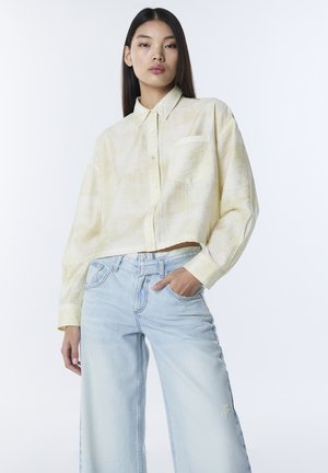 Przycięta żółta lniana koszula z przodem na guziki i kieszenią, zestawiona z jasnoniebieskimi jeansami o szerokich nogawkach z efektem sprania.