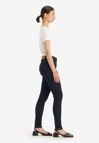 Sötétkék denim skinny farmer, közepes derékkal, diszkrét varrással és bőr logóval. Rövid ujjú fehér felsővel és fekete szandállal párosítva.