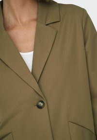 Blazer verde oliva con colletto a pattina, chiusura a un bottone e tessuto testurizzato. Presenta una tasca sul lato sinistro.