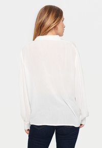 Blusa bianca leggera con maniche lunghe, dotata di un colletto semplice e polsini arricciati, vista da dietro. Tessuto liscio, vestibilità ampia.