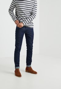 Chemise à manches longues rayée gris et marine, associée à un jean bleu foncé ajusté et des bottines en daim marron avec des accents bordeaux.