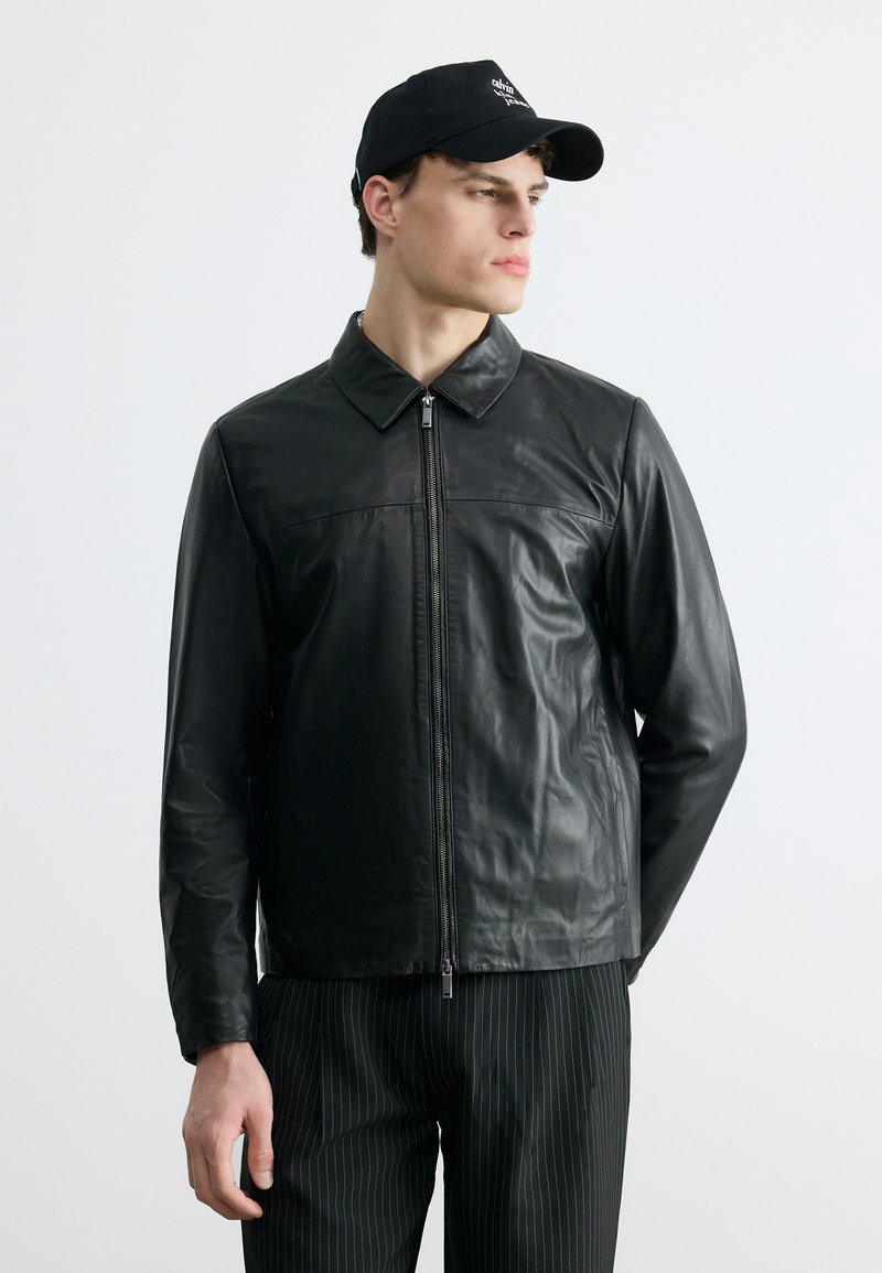 Νεαρός άνδρας που φοράει μαύρο δερμάτινο μπουφάν με φερμουάρ, παντελόνι με κάθετες ρίγες και μαύρο καπέλο μπέιζμπολ, κοιτάζει προς τα πλάγια μπροστά σε απλό φόντο.