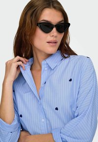 Šviesiai mėlyna maršelė su baltais dryžiais ir tamsiai mėlynomis širdelės siuvinėmis; apykaklė užsagstyta, rankovės suvirintos, derinama su juodais katės akies akiniais.