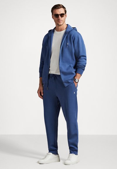 Μπλε φούτερ με κουκούλα και φερμουάρ, συνδυασμένο με ταιριαστά φόρμα, που έχει ελαστική μέση και λεπτομέρεια λογότυπου, συμπληρωμένο από λευκά αθλητικά παπούτσια.